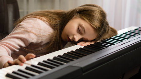 A girl rests her head on a piano keyboard, asleep during a practice session at home.の写真素材