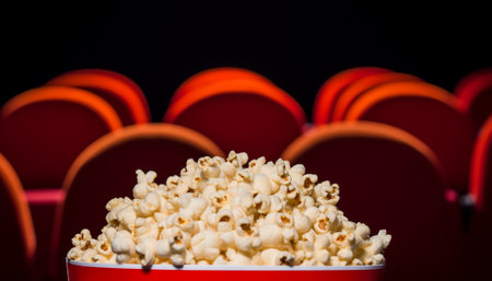 Overflowing popcorn in a red bucket with theater seats in the background.の素材