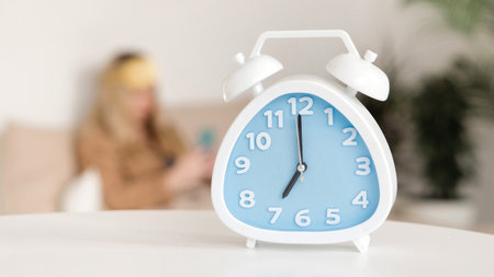 Alarm clock foreground, woman blurred in background. Morning scrolling news feed. The girl woke upの写真素材