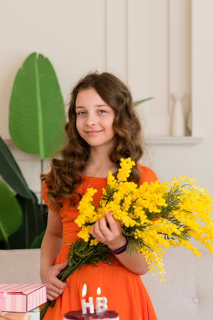 A young girl in an orange dress holding yellow flowers, smiling with birthday cake and gifts on the table. Portrait of a Beautiful Birthday Girlの写真素材