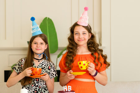 Two girls wearing birthday hats and holding colorful cups, celebrating indoors.の写真素材