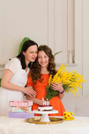 A young girl in an orange dress holding yellow flowers, standing next to a woman in a white dress,...