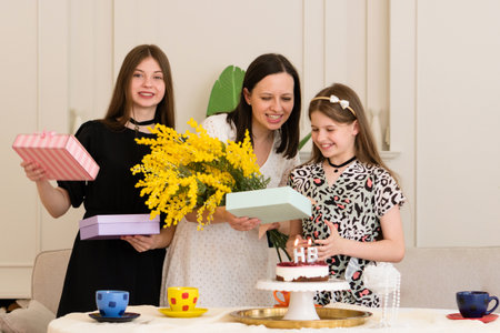 A woman and two girls celebrating a birthday with flowers, gifts, and a cake.の写真素材