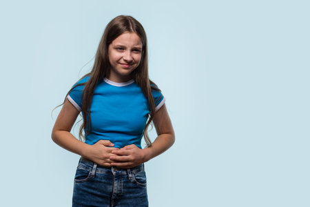 A young girl holding her stomach, showing signs of abdominal discomfort, standing in a studio setting.の写真素材