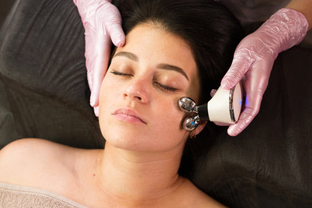 A beautician uses a microcurrent device on a client's cheek, performing a facial massage to enhance...