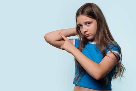 A young girl pointing at a cut on her elbow, looking worried, standing in a studio setting.の写真素材