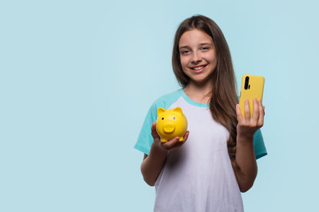 A young girl smiles as she holds a yellow piggy bank and a matching yellow smartphone, representing savings and modern financial management.の写真素材