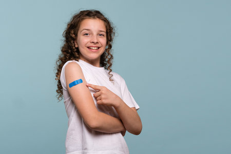Smiling girl pointing at a blue bandage on her arm after receiving a vaccine.の写真素材