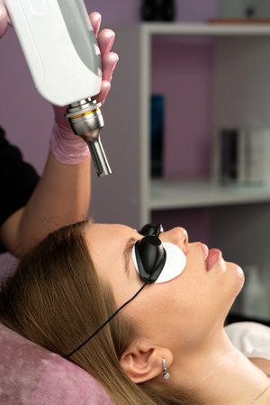 Esthetician aligns laser device over eyebrow for tattoo removal. Cosmetic medical procedure in a beauty salon. Hardware cosmetology treatmentの写真素材
