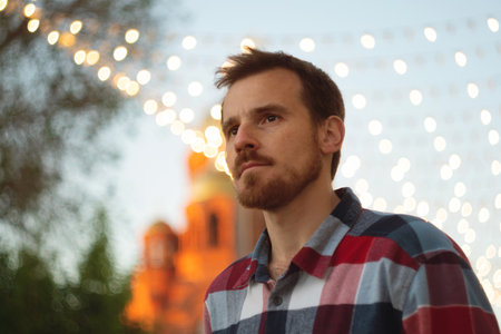 Thoughtful young man under festive outdoor city lights. Guy with beard and modern hairstyle wearing...