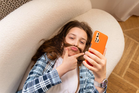 Teen girl in casual clothes taking playful selfie with hair mustache on smartphone. Spend free spare time in living room. Concept of lifestyle, use technologyの写真素材
