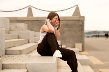 Young woman sitting on stone steps, laughing while talking on phone outdoors. Natural feel with soft...
