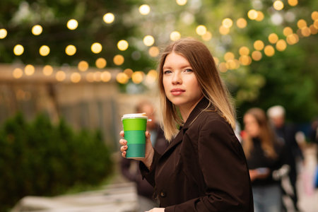 Young woman holding coffee cup walking outdoors casually. Great natural vibe and showcase stylish, relaxed mood. Outfit looks effortlessly chic.の写真素材