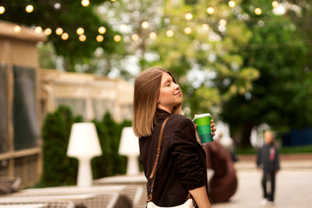 Young woman enjoying coffee outdoors near riverside promenade. Great natural vibe and showcase stylish, relaxed mood. Outfit looks effortlessly chic.の写真素材