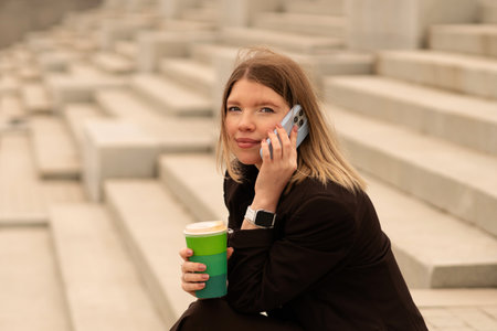 Young woman talking on phone and holding coffee cup outdoors. Natural feel with soft light. Casual, confident pose and expression add genuine and approachable vibe.の写真素材