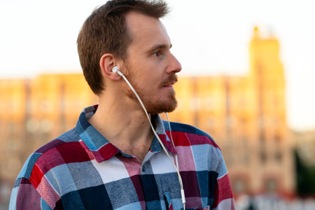 Profile of man listening to music outdoors at sunset. Guy with beard and modern hairstyle wearing in...