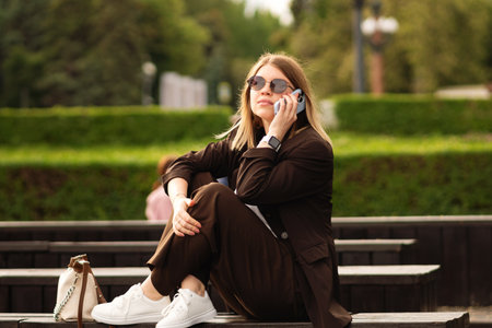 Young woman in brown suit sitting and using smartphone outdoors. Great natural vibe and showcase stylish, relaxed mood. Outfit looks effortlessly chic.の写真素材