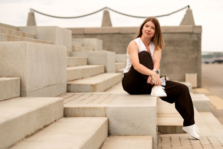Young woman sitting on steps, laughing while holding phone and looking down. Natural feel with soft light. Casual, confident pose and expression add genuine and approachable vibe.の写真素材