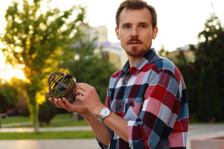 Young man holding antique globe thoughtfully during sunset outdoors. Astronomer holds an armillary sphere outside during an open class on spherical astronomyの写真素材
