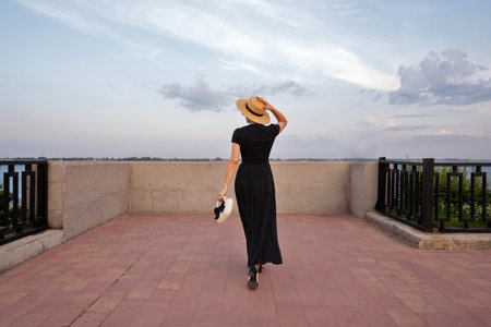 Woman in polka dot dress admires evening sky by river. An unrecognizable girl walks on the waterfront on a warm summer evening. View from behindの写真素材