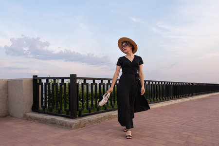 Stylish woman walks on promenade at sunset in black dressの写真素材