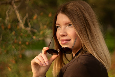 Young smiling woman holding sunglasses outdoors on sunny day.
