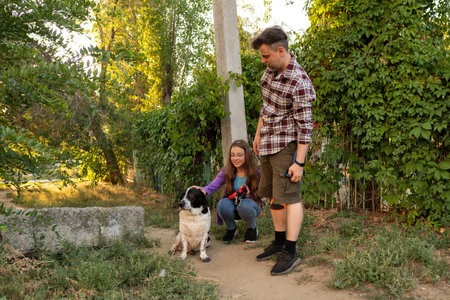 Man stands beside his young daughter crouching to pet their black and white dog on a dirt path,...