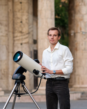 A man in a white shirt stands beside a mounted telescope in front of tall stone columns, posing...