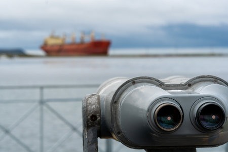 A weathered set of viewing binoculars in sharp focus with a blurred cargo ship in the background across the water under cloudy skies. Nevelsk, Sakhalin Islandの写真素材