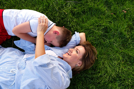 A joyful mother reclines on vivid grass, holding her drowsy son close as they share warmth and affection during a carefree summer afternoon in the park.の写真素材