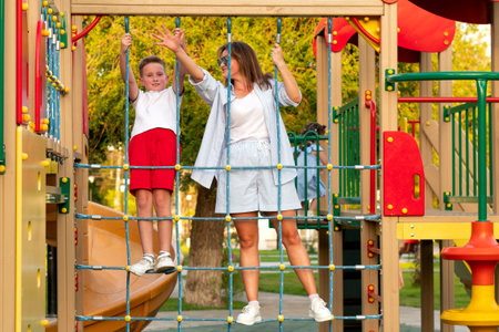 Smiling mother and her son grip rope grid bridge at colorful park playground, balancing together and waving during lively summer bonding time outdoorsの写真素材