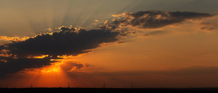 The orange sun sets behind clouds, with bright sun rays breaking through and illuminating the sky, silhouetted horizon and power lines visible in the distanceの写真素材