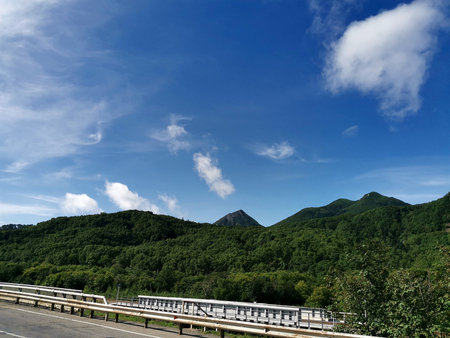 Lush forested mountains rise beneath a clear summer sky, while a roadside guardrail runs along the highway foreground, capturing a peaceful travel scene. Nature of Sakhalin Island.の写真素材