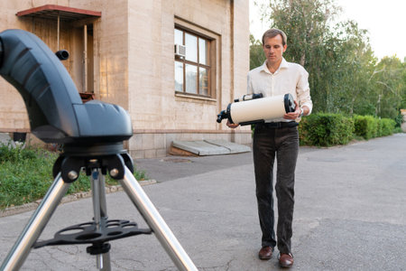 A man in dark trousers and white shirt holds a telescope tube while approaching a metal tripod setup...