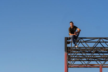 A male freerunner sits near the edge of a tall red steel parkour frame, arms folded and legs relaxed, preparing to descend from height in bright daylight.の写真素材