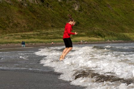 Girl in a red shirt leaps joyfully above the foamy sea waves near the beach, capturing a carefree seaside moment under the sun.  Tikhaya Bay in the Sea of ââOkhotsk, Sakhalin Islandの写真素材