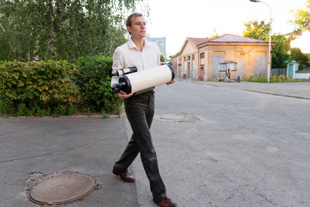 A man in formal clothes holds a large white telescope tube while walking across a paved urban area with background buildings, preparing for astronomical observation.の写真素材