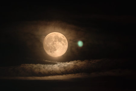Bright full moon glowing through thin clouds in a dark night sky. Atmospheric lunar light creates a...