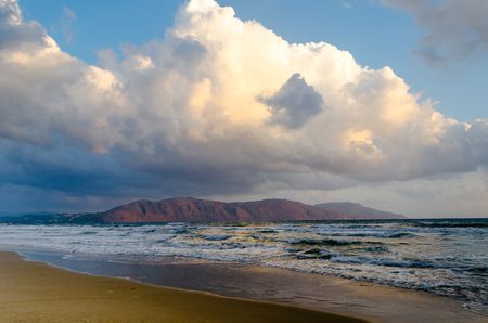 Huge cloud over the sea lit by the rising sun, blue waves, brown sand and red hillの写真素材