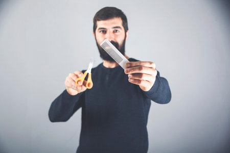 handsome man with long dark beard holding a hairbrush and scissors. hair and beard care in a hairdresser.の写真素材