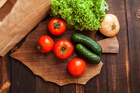 Tomatoes, cucumbers, lettuce, onions on a cutting board. Paper bag, vegetables, wooden brown background.の写真素材