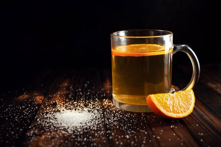 Tea with orange, sprinkled sugar, sliced orange, wooden brown background. Citrus drink in a transparent cup, empty space for text. Food composition on a black background in low key.の写真素材