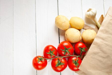 Beige paper bag on a wooden white background, vegetables from the store. Red tomatoes, yellow young potatoes, garlic. Empty space for text.の写真素材
