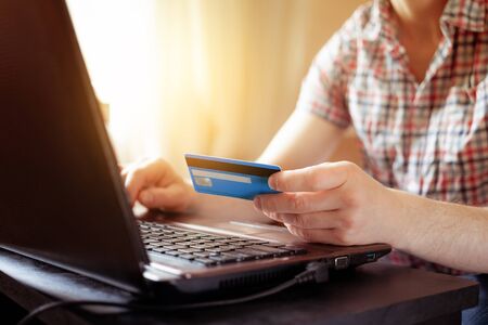 A man in a plaid shirt sits at a laptop and selects products in an online store. Payment by credit card. Home shopping. Online shopping on the Internet.の写真素材