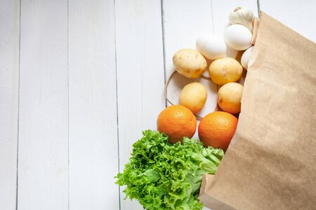 Beige paper bag on a wooden white background, vegetables from the store. Salad, oranges, eggs, garlic, potatoes. Empty space for text.の写真素材