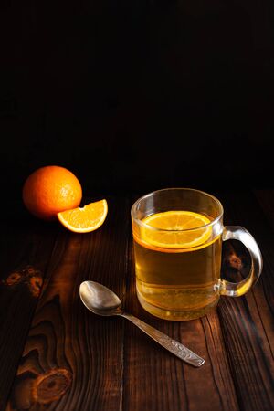 Tea with orange, metal spoon, sliced orange, wooden brown background. Citrus drink in a transparent cup, empty space for text. Food composition on a black background in low key.の写真素材