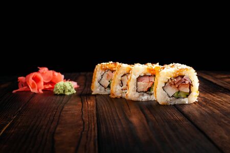 Rolls lie on a wooden brown board. Black background. Green wasabi, pickled ginger slide. Warm fried rolls with bacon and smoked chicken.の写真素材