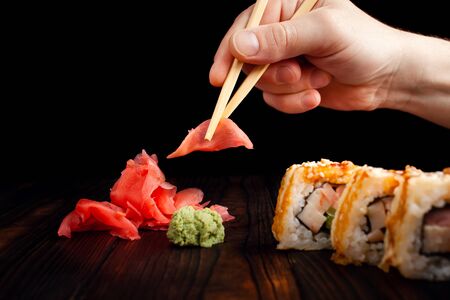 A man takes wooden sticks to ginger with a wooden board. Black background. Green wasabi, pickled ginger slide. Warm rolls with bacon and chicken.の写真素材