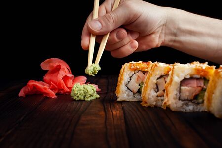 A man takes wooden sticks to wasabi with a wooden board. Black background. Green wasabi, pickled ginger slide. Warm rolls with bacon and chicken.の写真素材