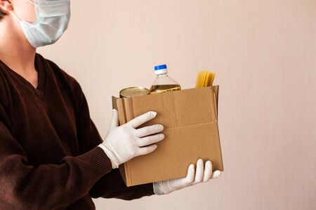 A volunteer delivers food to those in need. Delivery donation. Sunflower oil, canned food, pasta in a box. Hands in gloves, masked man.の写真素材